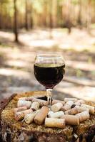 A glass with a red wine and wine corks on a stump on a background of a summer forest in a sunny day. photo