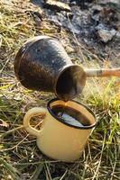 taza de metal amarillo con café caliente y cezve sobre un césped al fondo de la hoguera. foto