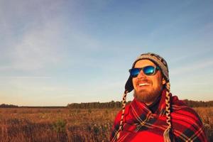 un joven sonriente feliz con gorro de punto, gafas de sol y manta se encuentra con el amanecer en el fondo de un campo y un cielo azul. tonificado foto