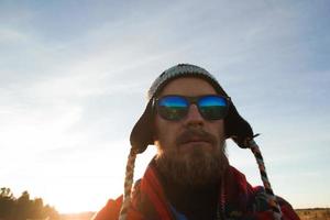 joven feliz con gorro de punto, gafas de sol y manta en el fondo de un campo y cielo azul. foto