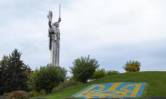 Statue of the Motherland against the blue sky. Coat of arms of Ukraine on the lawn, lined with blue and yellow stones. The trident, the official symbol of the state. Ukraine, Kyiv - October 08, 2022. photo