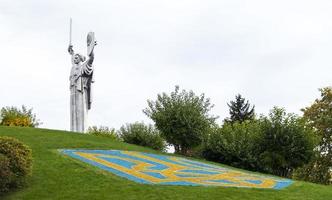 Statue of the Motherland against the blue sky. Coat of arms of Ukraine on the lawn, lined with blue and yellow stones. The trident, the official symbol of the state. Ukraine, Kyiv - October 08, 2022. photo