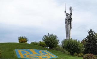 Statue of the Motherland against the blue sky. Coat of arms of Ukraine on the lawn, lined with blue and yellow stones. The trident, the official symbol of the state. Ukraine, Kyiv - October 08, 2022. photo