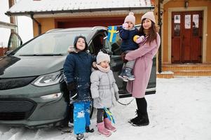 Young swedish mother with kids hold Sweden flags and charging electric car in the yard of her house at winter. photo