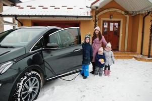 Young woman with kids hold eco bags and charging electric car in the yard of her house . photo