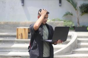 Attractive young Asian man using laptop confused in co-working space with unhappy face photo