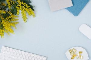 sobre un fondo plano azul con una flor de mimosa amarilla, bloc de notas de teclado y clips de papelería, un escritorio floral femenino. el concepto de una elegante mañana de primavera. vista superior y espacio de copia foto