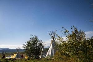 View of teepees on field amidst trees with blue sky in background on sunny day photo
