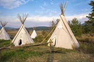 Teepees on field against trees with blue sky in background on sunny day photo