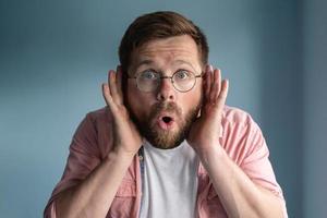 Man with glasses eavesdrops with his hands to ears, he opened mouth in shock and looks with big eyes. photo