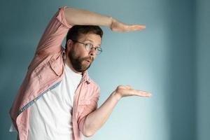 Man in glasses shows the size of something with hands, standing against a blue background. Copy Space. Place for an advertisement. photo