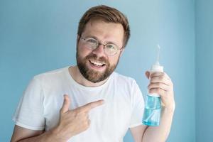 Cute smiling man holds a portable irrigator in hand, he recommends this oral hygiene product and points to it with his finger photo