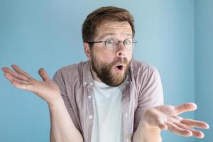 Bearded man in glasses made an innocent surprised expression and makes a bewildered gesture with hands. Studio, blue background. photo