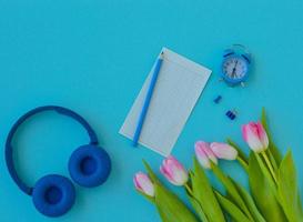 Flat lay home office desk. Workspace with pink tulips bouquet, headphones, alarm clock, pencil and a piece of paper for notes on blue background. Top view feminine background.