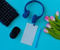 Flat lay home office desk. Workspace with laptop, pink tulips bouquet, headphones, pencil and a piece of paper for notes on blue background. Top view feminine background. photo