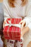 Happy young woman holding a present surrounded by gift boxes sits cross-legged on a camel couch. Christmas gift in the hands of a girl, close-up. photo