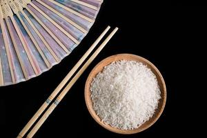 arroz blanco crudo, abanico de mano y palillos sobre fondo negro. arroz largo sin cocer en un plato de madera. foto