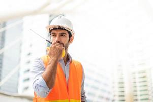 el ingeniero caucásico usa un walkie-talkie para hablar, usa chaleco naranja y un gran sombrero duro en el sitio de trabajo del centro de la ciudad. foto