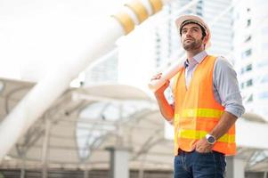 Young caucasioan man holding a big paper, guy wearing light blue shirt and jeans with orange vest and white helmet for security in construction area. photo