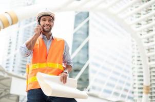 el ingeniero caucásico usa un teléfono inteligente para hablar, usa un chaleco naranja y un gran sombrero duro, y la otra mano sostiene el plano blanco en el sitio de trabajo del centro de la ciudad. foto