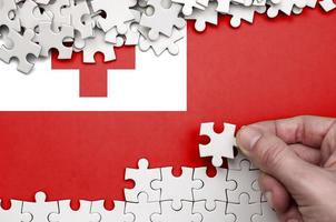 Tonga flag is depicted on a table on which the human hand folds a puzzle of white color photo