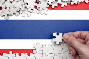 Thailand flag is depicted on a table on which the human hand folds a puzzle of white color photo