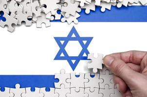 Israel flag is depicted on a table on which the human hand folds a puzzle of white color photo