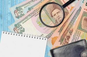 100 billetes de hryvnias ucranianos y lupa con monedero negro y bloc de notas. concepto de dinero falso. busque diferencias en los detalles de los billetes de dinero para detectar falsos foto