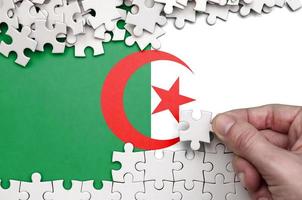 Algeria flag is depicted on a table on which the human hand folds a puzzle of white color photo