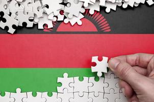 Malawi flag is depicted on a table on which the human hand folds a puzzle of white color photo