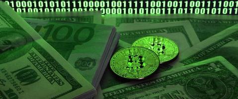 Two bitcoins lies on a pile of dollar bills on the background of a monitor depicting a binary code of bright green zeros and one units on a black background. Low key lighting photo