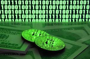 Two bitcoins lies on a pile of dollar bills on the background of a monitor depicting a binary code of bright green zeros and one units on a black background. Low key lighting photo