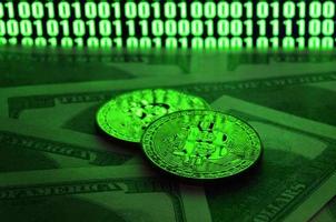 Two bitcoins lies on a pile of dollar bills on the background of a monitor depicting a binary code of bright green zeros and one units on a black background. Low key lighting photo