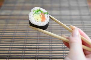 A hand with chopsticks holds a sushi roll on a bamboo straw serwing mat background. Traditional Asian food photo