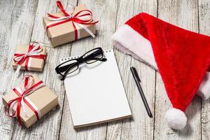 Christmas and Business Items with Copy Space Santa Cap Notepad Pen Glasses and decorated Gift Boxes photo