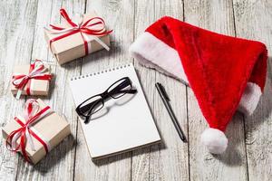 Christmas and Business Items with Copy Space Santa Cap Notepad Pen Glasses and decorated Gift Boxes photo