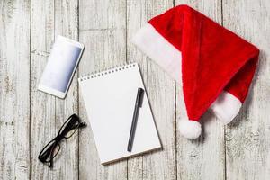 Christmas and New Year background with smartphone, red Santa hat, glasses, notepad and pen for your wishes photo