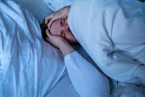 Young man lies in bed with a severe headache, he clasps head with hands and makes a grimace of suffering. photo