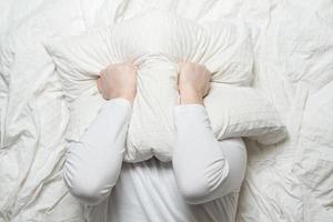 Man in white pajamas cannot sleep because of the noise and, in stress, covers head with a pillow. Insomnia concept. Top view. photo