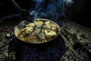 Large chunks of freshly caught fish are fried in a pan over a campfire, over coals, in the evening, at a camping. Lifestyle. photo