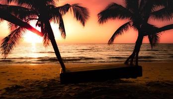 silueta de palmera y columpio de madera en la playa durante la puesta de sol de una hermosa playa tropical sobre fondo de cielo rosa foto