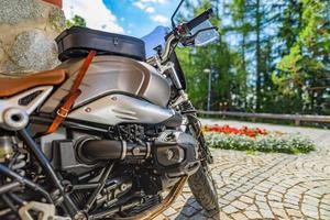 Closeup motorcycle parked on driveway road in warm sunny light. Blurred forest trees, park flowers and gravel stone road. Classic vintage motorbike, outdoor sport recreational pursuit. Nature travel photo