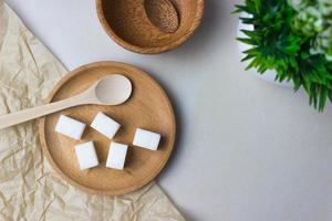 Sugar cubes in a wooden plate with a spoon on the kitchen table with utensils and green plant. Top view photo