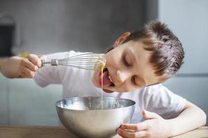 niño cocinando en la cocina. niño revolviendo masa para un pastel en el recipiente de acero. el niño prueba la comida y lame un batidor