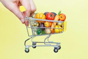 carrito de compras con frutas y verduras en la mano de las personas sobre fondo amarillo. entrega de alimentos concepto de compras por internet foto