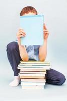 Schoolboy is sitting with a stack of books and reading and covers his face with a book isolated on blue background