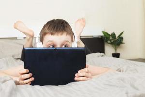 child boy lying on bed and covered his face with a pc tablet with wireless headphones photo
