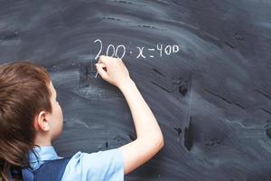 Boy standing back in front of school blackboard and writing. Schoolboy solves math example at the chalkkboard photo