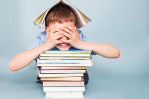 colegial sentado con un montón de libros escolares y cubre su cara con las manos aisladas en un fondo azul foto