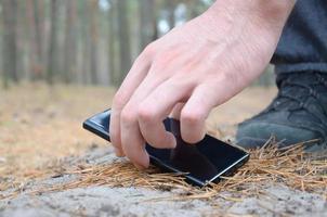 mano masculina recogiendo el teléfono móvil perdido de un suelo en el camino de madera de abeto de otoño. el concepto de encontrar algo valioso y buena suerte foto